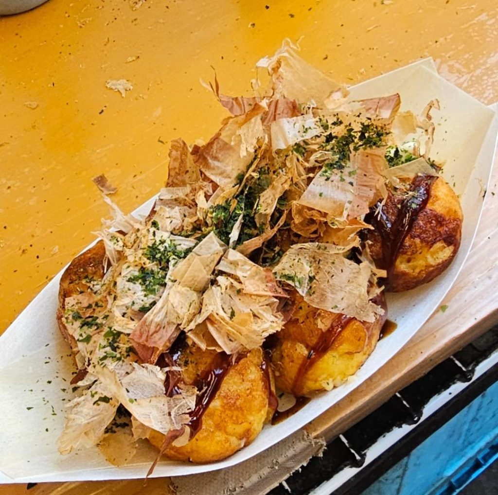 Close-up of takoyaki served in a paper boat, generously topped with bonito flakes, seaweed powder, and sauce, taken at Ueno Ameyoko.