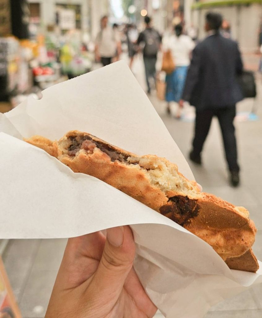 A partially eaten taiyaki revealing sweet red bean paste filling, held by hand with a blurred bustling street view of Kichijoji in the background.